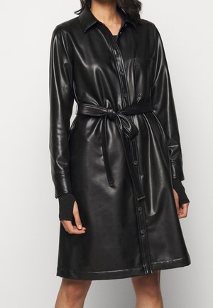 Vestido camisa de cuero negro con cuello, bolsillos frontales y cintura ajustada con un lazo. Cuenta con mangas largas con puños acanalados y botones en la parte delantera.