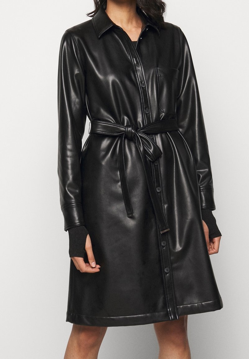 Černé kožené šaty košile s límečkem, předními kapsami a zavazováním v pase. Mají dlouhé rukávy s žebrovanými manžetami a knoflíky po celé délce.