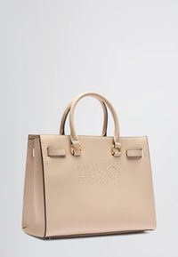 Sac à main synthétique beige avec deux poignées arrondies, logo en relief, quincaillerie dorée, design structuré et garniture sombre contrastante.
