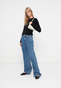 Crna pletena majica, široke plave traperice i crne balerinke. Model drži bijelu torbicu. Jednostavna pozadina pojačava fokus na detalje odjevne kombinacije.