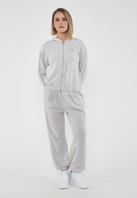 Tuta grigio chiaro con giacca con zip e pantaloni coordinati. Presenta polsini e vita elasticizzati, con dettaglio del logo sulla giacca. Sneakers bianche.