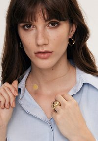 Złote kolczyki w kształcie kół, delikatna złota naszyjnik z okrągłym wisiorkiem i złoty pierścionek o rzeźbiarskiej formie. Modelka ma na sobie jasnoniebieską koszulę.