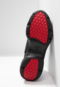 Schwarze Schuhsohle mit einem strukturierten Muster, das rote Akzente und ein Logo in der Mitte aufweist. Der Rand ist gummiert für besseren Halt und Langlebigkeit.