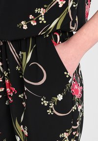 Svart blommig jumpsuit med röda och vita blommor, gröna blad och beige detaljer. Inkluderar en sidoficka för bekvämlighet.