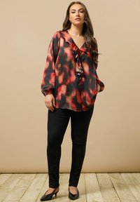 Kvinde iført en rød og sort bluse med volanger, sorte bukser og sorte hæle, der står på et trægulv foran en beige baggrund.