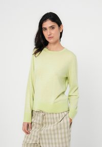 Ανοιχτό πράσινο πουλόβερ με μακριά μανίκια και στρογγυλή λαιμόκοψη, απαλή υφή υφάσματος και ριμπ τελειώματα στα μανίκια και στο τελείωμα, σε συνδυασμό με καρό σορτς.