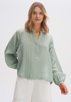 Blusa verde chiaro a nido d'ape con maniche lunghe e voluminose, un'apertura centrale con bottoni e una vestibilità ampia. Abbinata a pantaloni corti bianchi.