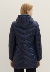 Marinblå pufferjacka med huva, chevronmönster och glänsande yta. Elastiska muddarna och den medellånga designen ökar värmen och skyddet.