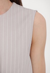 Haut sans manches gris clair avec un motif en chevrons texturé et de fines rayures blanches verticales. Tissu lisse avec une finition douce.