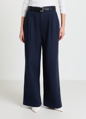 Pantalon large bleu marine avec plis sur le devant, taille haute et ceinture noire. Associé à des chaussures blanches à bout pointu. Texture de tissu lisse.