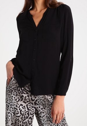 Mujer vistiendo una camisa negra de manga larga con botones y pantalones de talle alto con estampado de leopardo en gris y negro. Una mano en el bolsillo.