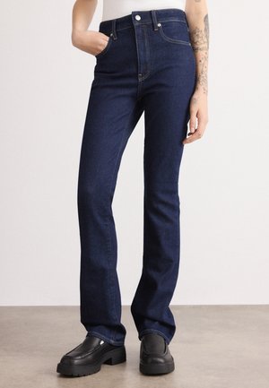 Persoon die donkerblauwe recht gesneden spijkerbroek draagt, zwarte chunky loafers, met één hand in de zak en zichtbare armtattoos.