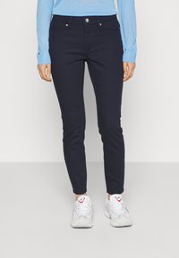 Vaqueros elásticos de color azul marino con un ajuste slim, cinco bolsillos y detalles en botón plateado, combinados con zapatillas blancas con acentos coloridos.