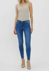 Vaqueros ajustados de mezclilla azul con una cintura de media altura, bolsillos frontales y una textura suave, combinados con una blusa sin mangas de color gris claro y tacones beige.