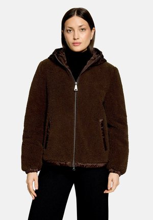 Frau trägt eine dunkelbraune Fleecejacke mit Reißverschluss, Kapuze, elastischen Bündchen und Seitentaschen über einem schwarzen Rollkragenpullover und schwarzen Hosen.