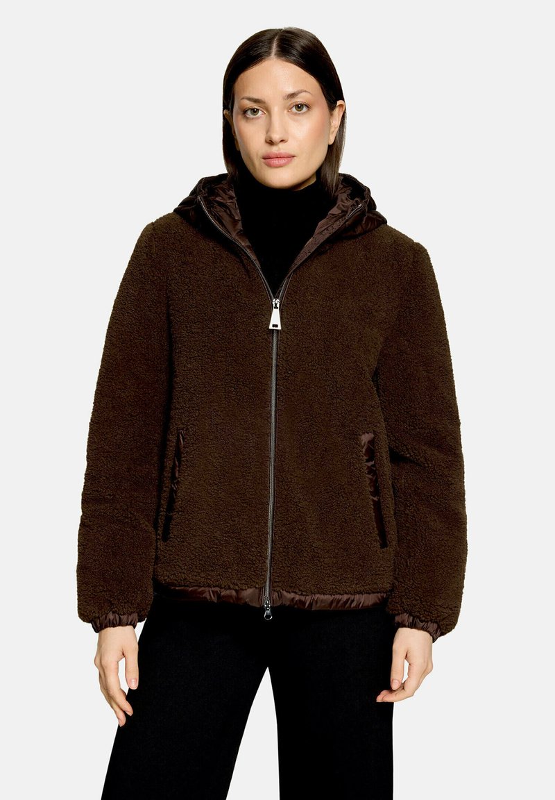 Femme portant une veste polaire marron foncé à fermeture éclair avec capuche, poignets élastiques et poches latérales, par-dessus un col roulé noir et un pantalon noir.