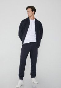 Marineblaue Kapuzenjacke mit Reißverschluss und Kordelzug; kombiniert mit passenden Jogginghosen. Ein weißes Rundhalsausschnitt-T-Shirt und weiße Sneakers vervollständigen den Look.