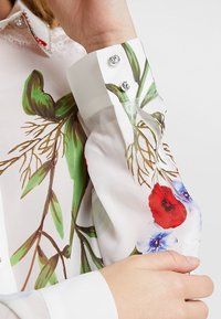 Blouse fleurie avec une base blanche ornée de motifs floraux rouges et bleus, manches longues, col en dentelle et boutons argentés.