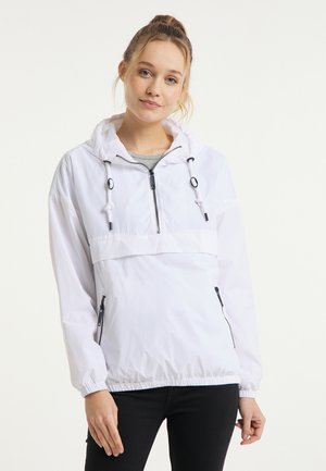 Junge Frau trägt eine weiße winddichte Jacke mit Kapuze und Frontreißverschluss sowie schwarze Hose, steht vor einem einfarbigen hellen Hintergrund.