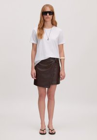 Weißes Kurzarm-T-Shirt, brauner Lederwickelrock, schwarze Flip-Flops und Sonnenbrille. Bemerkenswertes Knotendetail am Rock. Einfaches Design.