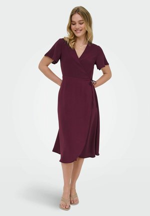 Ung kvinde med blondt hår iført en kortærmet, knælængde dyb lilla slå-om-kjole og beige åbne tå-sko, smilende med hænderne bag ryggen.