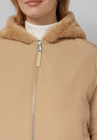 Beige jack met een nepbont capuchon, voorzien van een zilveren ritsluiting en een zachte textuur. Eenvoudig ontwerp zonder extra versieringen.