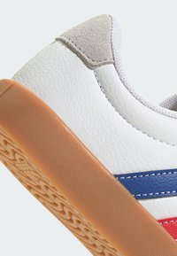 Zapatilla de cuero blanca con rayas azules y rojas, acento de ante gris y suela de goma texturizada. Los detalles de costura son visibles a lo largo de las costuras.