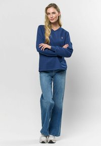 Sieviete ar gariem gaišiem matiem, stāvot ar saliktām rokām, tērpusies ziliem džemperiem, platiem džinsiem un baltām sporta apaviem, uz vienkārša fona.