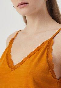 Oransje camisole med V-hals og bølget blondekant, samt justerbare tynne stropper. Mykt stoff med glatt tekstur.