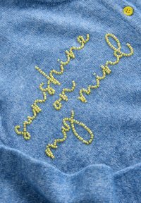 Blauer Pullover mit gelber Perlenstickerei, auf der "sonnenschein im Kopf" steht. Weiche Textur mit lässiger Passform und einem gelben Knopfakzent.