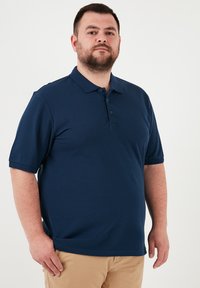 Tamsiai mėlynas polo marškinėliai, pagaminti iš lygios medžiagos, su trumpomis rankovėmis, trimis mygtukais ir laisvu kirpimu. Dėvimi su chaki spalvos kelnais.
