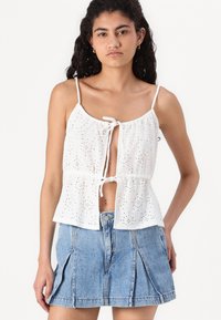 Witte katoenen tanktop met oogjespatroon, gerimpelde taille en veter sluiting. Gepaard met een lichtblauwe denimrok met plooien.