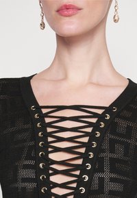 Femme portant un haut noir à lacets avec œillets dorés et boucles d'oreilles dorées pendantes, montrant le cou et le bas du visage.