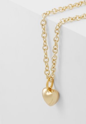 Collier chaîne en or avec un pendentif lisse en forme de cœur gravé d'un nom de marque, posé sur une surface géométrique blanche.