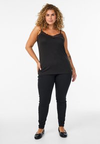Mujer con cabello rizado que lleva una camiseta de tirantes negra con encaje, pantalones negros y zapatos planos negros, de pie con una mano en la cadera frente a un fondo blanco.