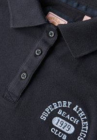 Camisa polo negra de tela texturizada, con cuello clásico, tapeta de tres botones y logo bordado en azul claro.