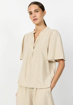Junge Frau mit dunklen, zurückgebundenen Haaren, die ein lockeres beige Kurzarmoberteil und passende Hose trägt, steht mit einer Hand in der Tasche.