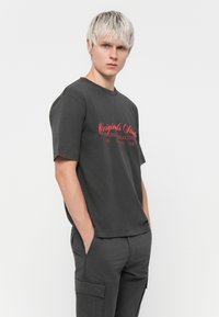 Tricou din bumbac gri închis, cu mâneci scurte și guler rotund, având un design cu text roșu inscripționat "Originals" pe partea din față.