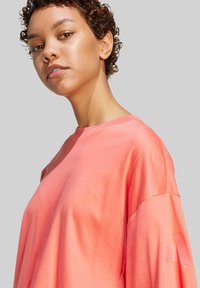 Korálová oversized T-shirt s voľným strihom, mäkký bavlnený materiál, krátke rukávy a výrazný výrez na ľavom rukáve.