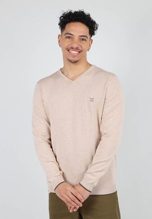 Pull beige à col en V en matière douce, avec des manches longues et des poignets côtelés avec une finition en vert foncé contrastant, porté sur un pantalon olive.