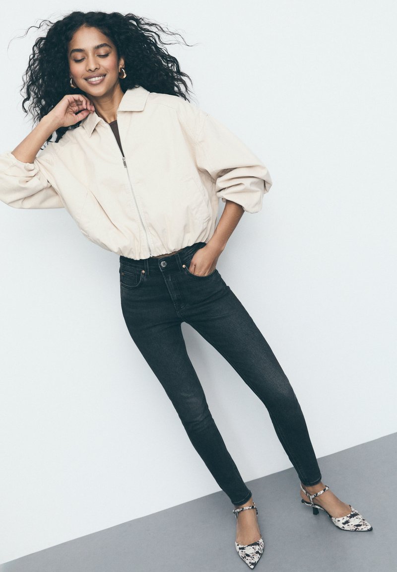 Beige cropped jasje met rits aan de voorkant, zwarte high-waisted skinny jeans en geprinte hakken met een spitse neus en kleine hak.