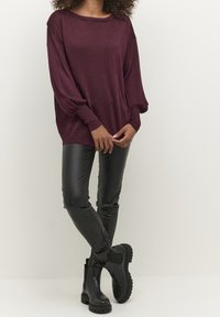 Bordeaux lopend vest met lange mouwen, gecombineerd met zwarte nepleer leggings en zwarte enkellaarsjes met gestructureerde zolen.