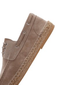Zapato de ante marrón claro con suela de yute trenzado, ojal metálico y diseño de cordones. Su forma es casual y redondeada.