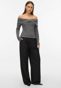 Kobieta w szarym, długim swetrze z odkrytymi ramionami i czarnych, szerokich spodniach, stoi z jedną ręką w kieszeni.