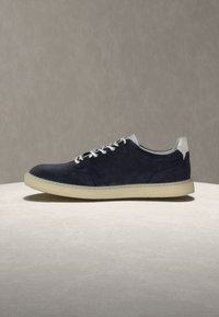 Baskets en daim bleu foncé avec lacets blancs, languette blanche au talon et semelle beige sur une surface texturée claire, sur fond gris.