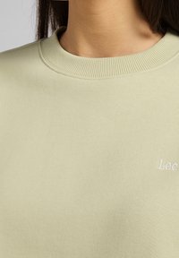 Sudadera verde claro con cuello redondo acanalado y logo bordado sutil. Textura suave con un acabado liso.