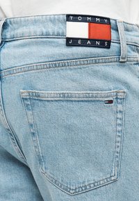 Φωτεινό γαλάζιο τζιν παντελόνι με πίσω τσέπη και υφαντή ετικέτα με το όνομα της μάρκας "TOMMY JEANS" μαζί με κόκκινες και λευκές λεπτομέρειες.