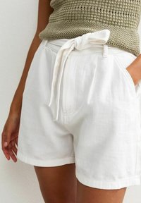 Shorts en lin blanc avec un détail de taille nouée, des poches avant et une coupe décontractée. Le tissu a une texture lisse et un subtil éclat.