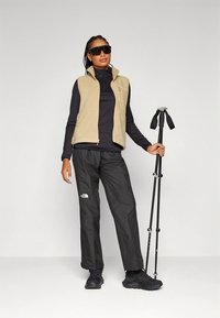 Beige fleecevest over en svart langermet skjorte, kombinert med svarte turbukser og mørke tursko. Holder en turstav.