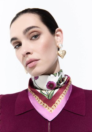 Mujer con cabello oscuro que lleva pendientes dorados en forma de corazón y una bufanda rosa con estampado floral sobre una camiseta burdeos.